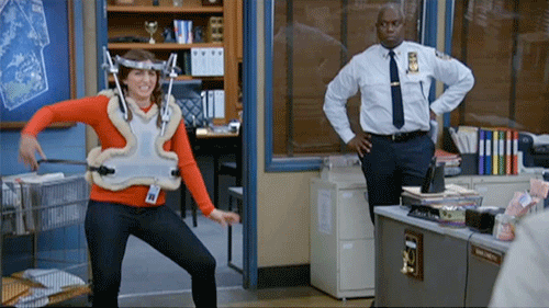GIF of Gina Linetti (Brooklyn 99) in an upper body medical brace, dancing in pain, watched by Captain Holt and the rest of the 99.